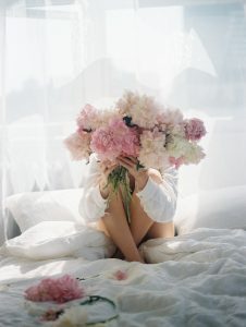 Woman sat on bed holding big bunch of flowers in front of her face