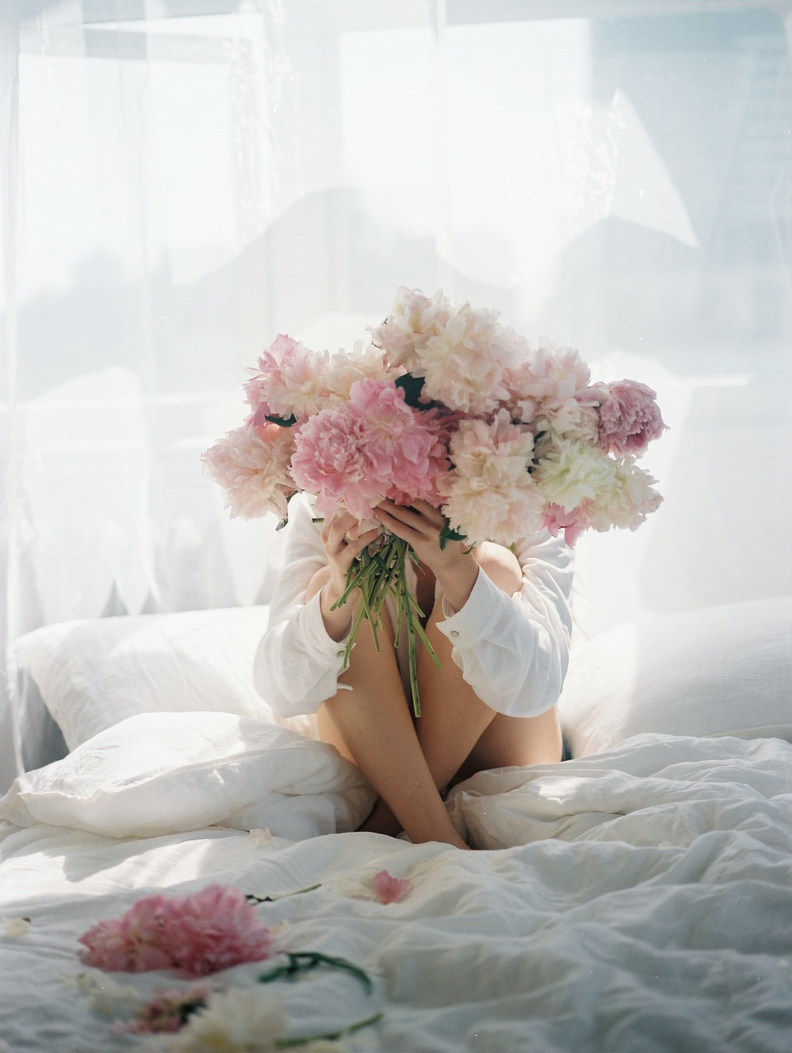 Woman sat on bed holding big bunch of flowers in front of her face