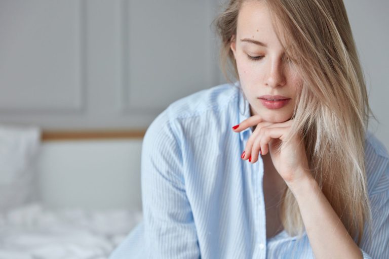 young blonde woman in light blue shirt sat on bed thinking