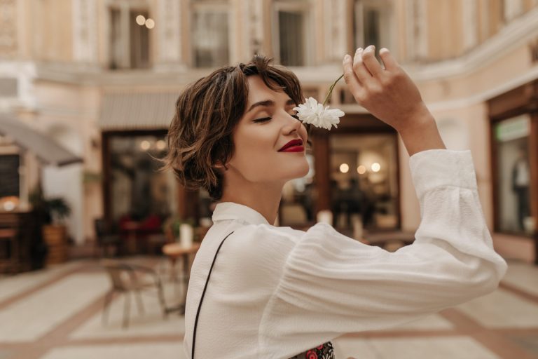brunette woman in white shirt smelling a white flower